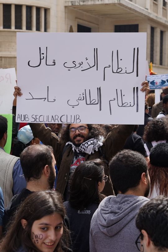 Man from the AUB Secular Club holding an Arabic sign from the 2019 Women's March in Beirut reads "The patriarchal system is a killer, the sectarian system is corrupt." (Laudy Issa)