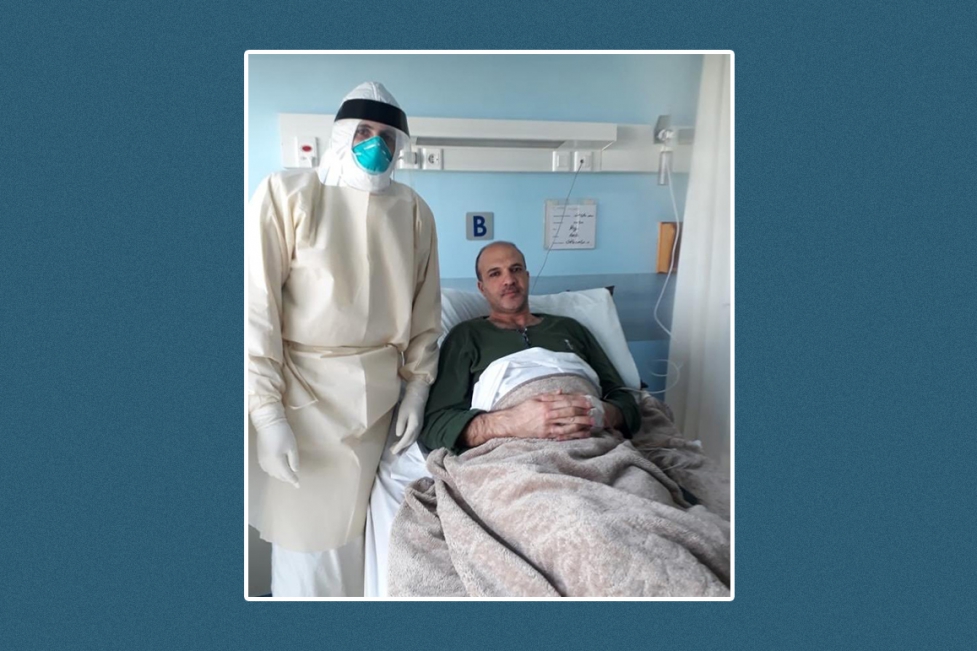 Health minister Hamad Hassan sits in a hospital bed at St. George Hospital while a healthcare worker fully cladded in a hazmat suit stands beside him.