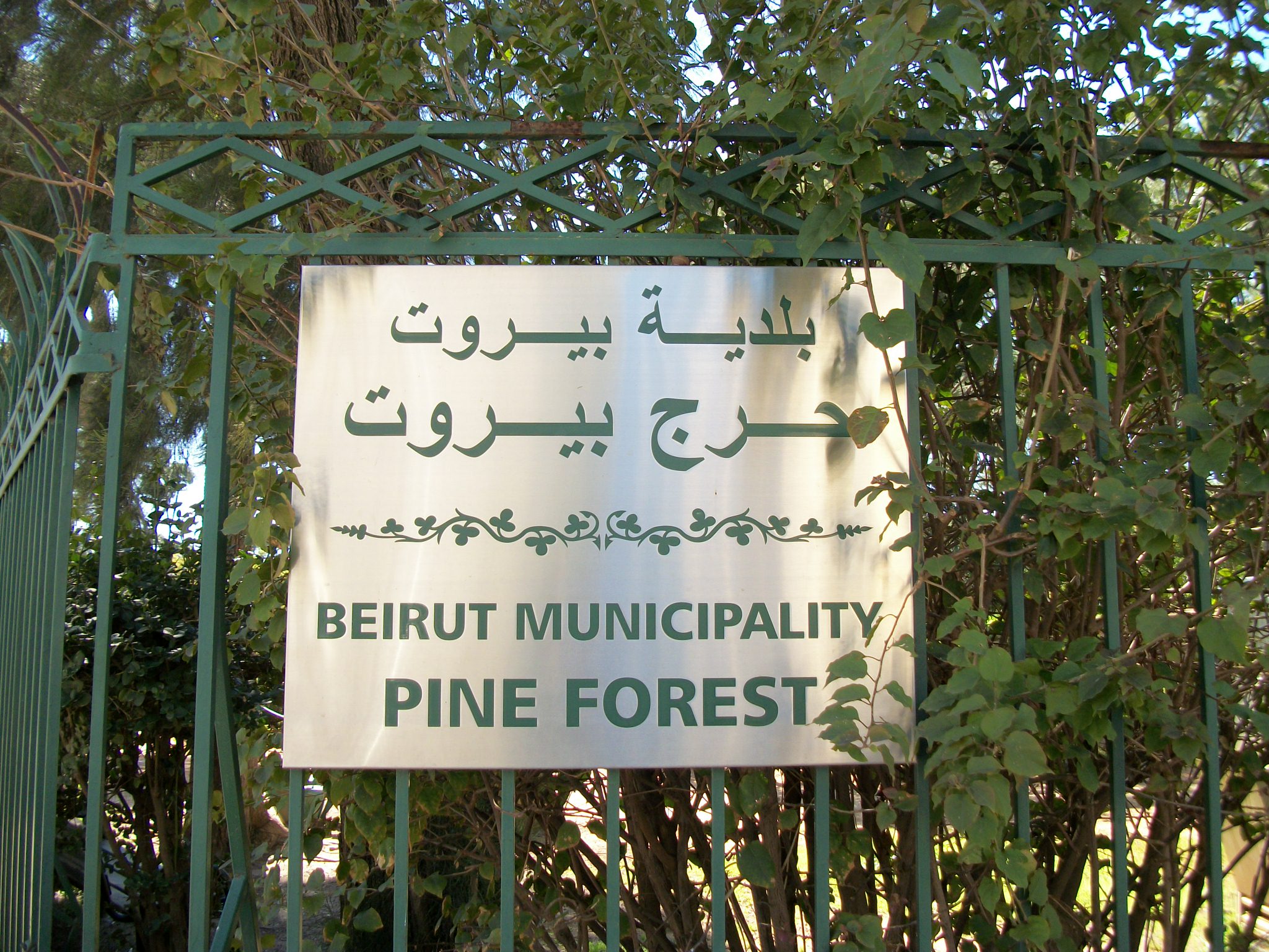 Photo showing a Beirut Municipality Horsh Beirut sign hung up on a green gate against a backdrop of trees