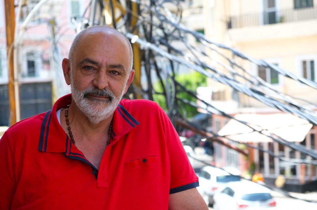 Tony, a man with a grey beard, smiles softly at the camera in Gemmayze. He is wearing a red shirt, and the background is blurred.