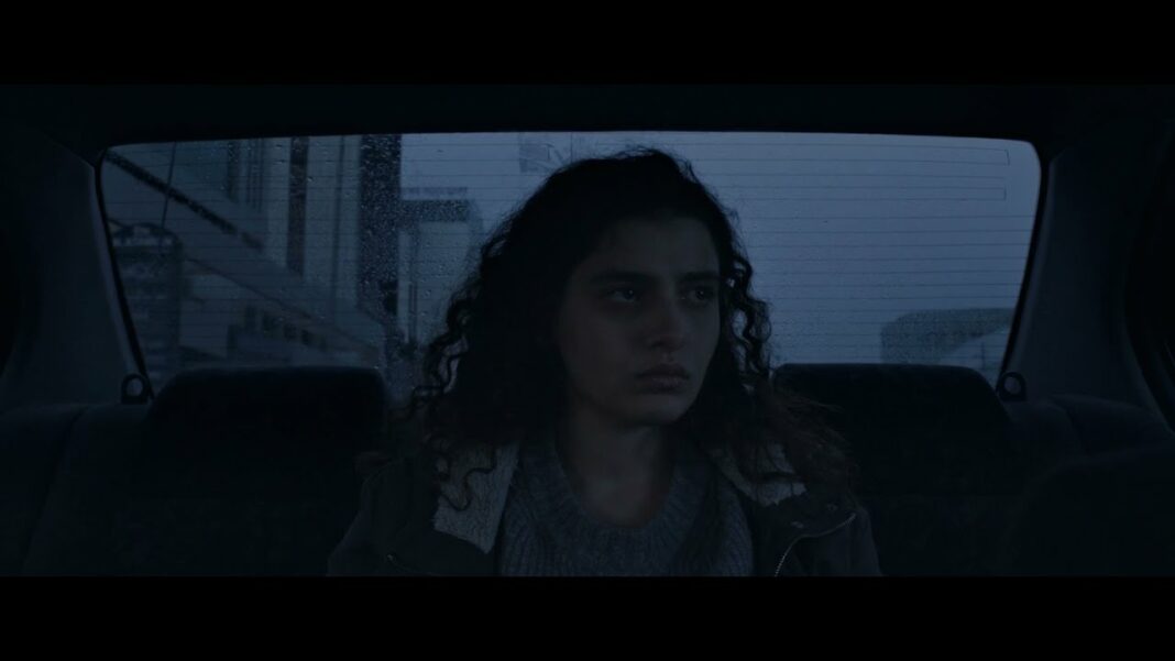 Manal Issa, who plays Jana in The Sea Ahead, sits in the back of a car looking out the window. She is centered in the middle of the image, whose colors and desaturated and dark. Her face is expressionless.