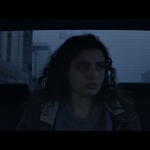 Manal Issa, who plays Jana in The Sea Ahead, sits in the back of a car looking out the window. She is centered in the middle of the image, whose colors and desaturated and dark. Her face is expressionless.