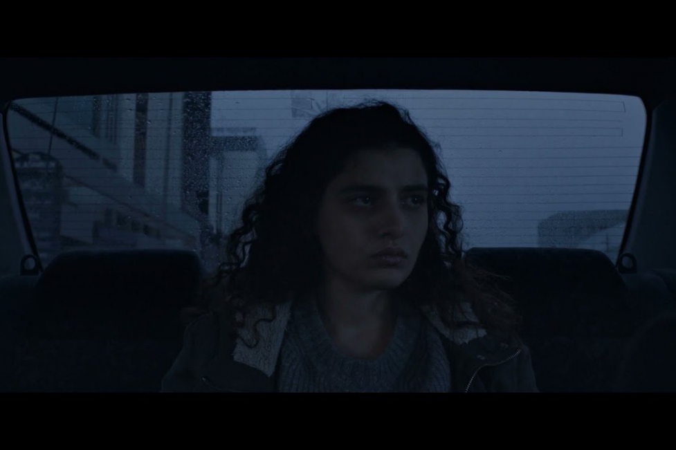 Manal Issa, who plays Jana in The Sea Ahead, sits in the back of a car looking out the window. She is centered in the middle of the image, whose colors and desaturated and dark. Her face is expressionless.